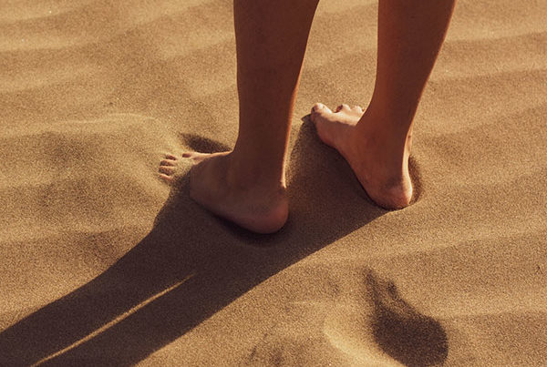 Close up of feet standing in dry sand, possibly of beach. Sun casts strong shadow of feet and legs to left.