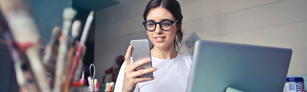 Pretty young woman in glasses using a mobile phone and credit card in front of open laptop