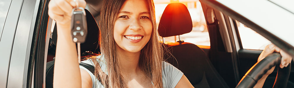 Smiling young woman looks at camera. She is seated in the driver's seat of a car and proudly holding up the keys to her new car.