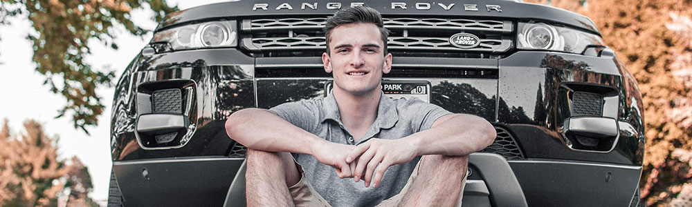 Smiling young man in shorts, sits in front of his Land Rover Evoque, autumn leaves in background