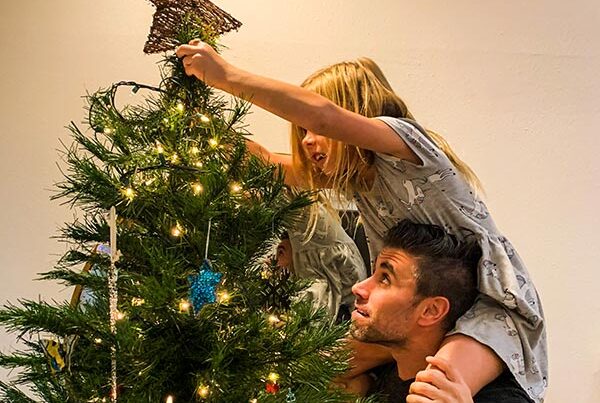 Dad lifts blonde daughter on his shoulders to place golden star on the top of their family christmas tree.