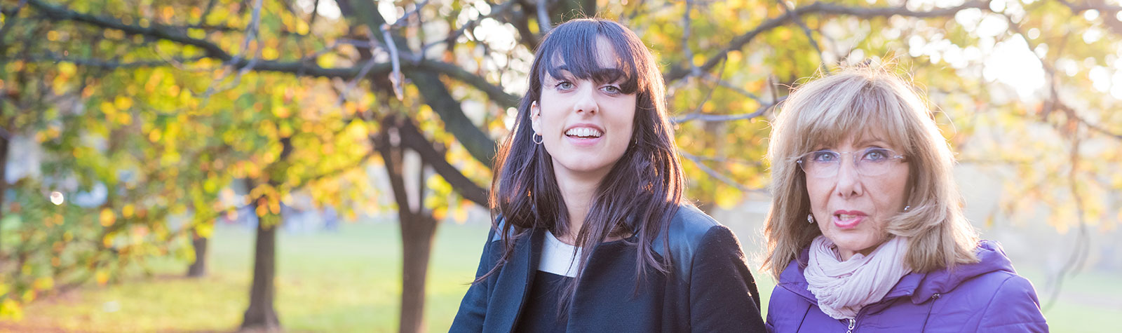 Mother and adult daughter walking together in the park they travelled to using day insurance. Sunny day, smiling ladies. Trees with autumn leaves